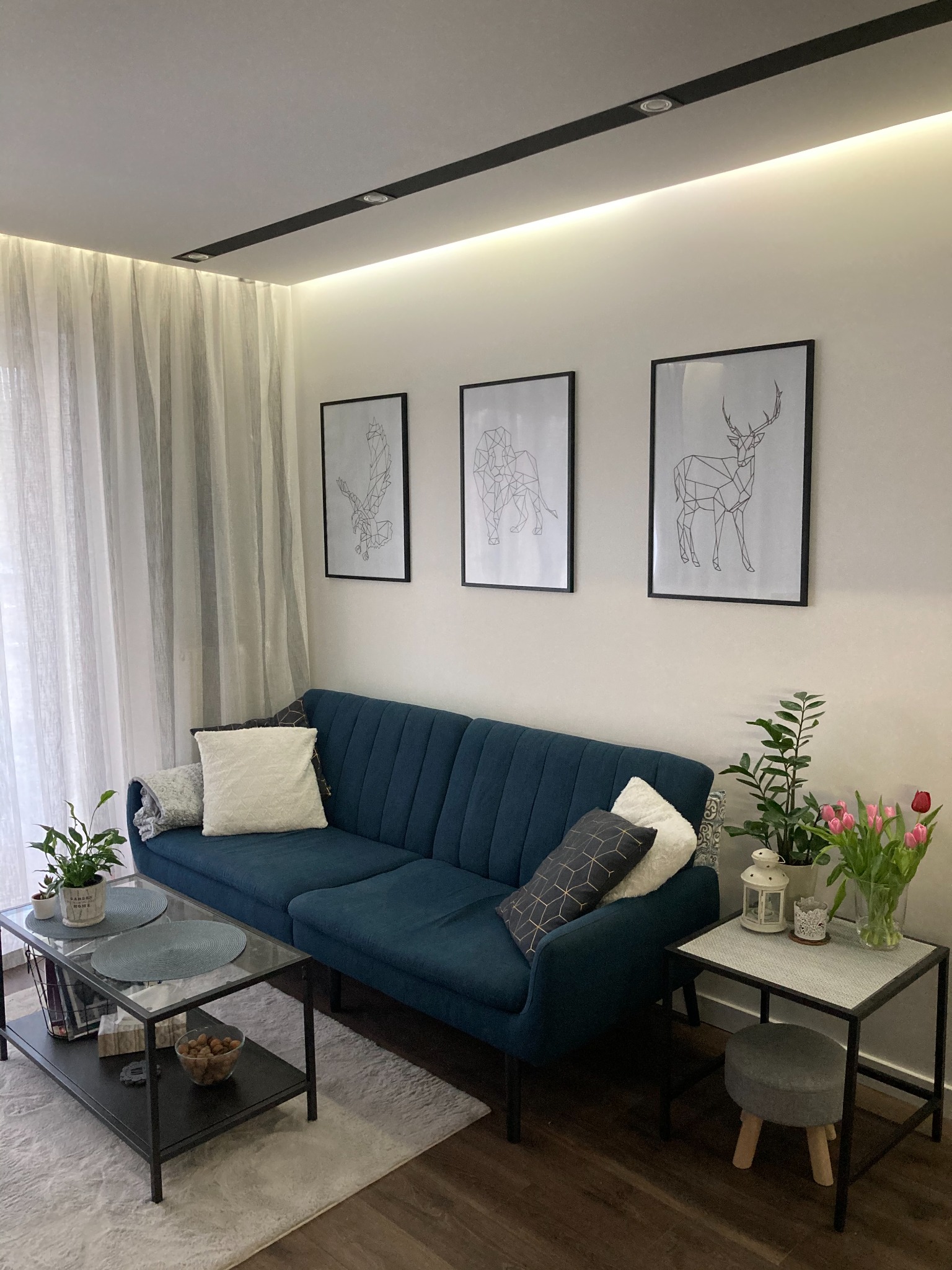 Nowoczesny salon z turkusową sofą, geometrycznymi obrazami na ścianie, szarymi zasłonami i stolikami kawowymi z roślinami i tulipanami.