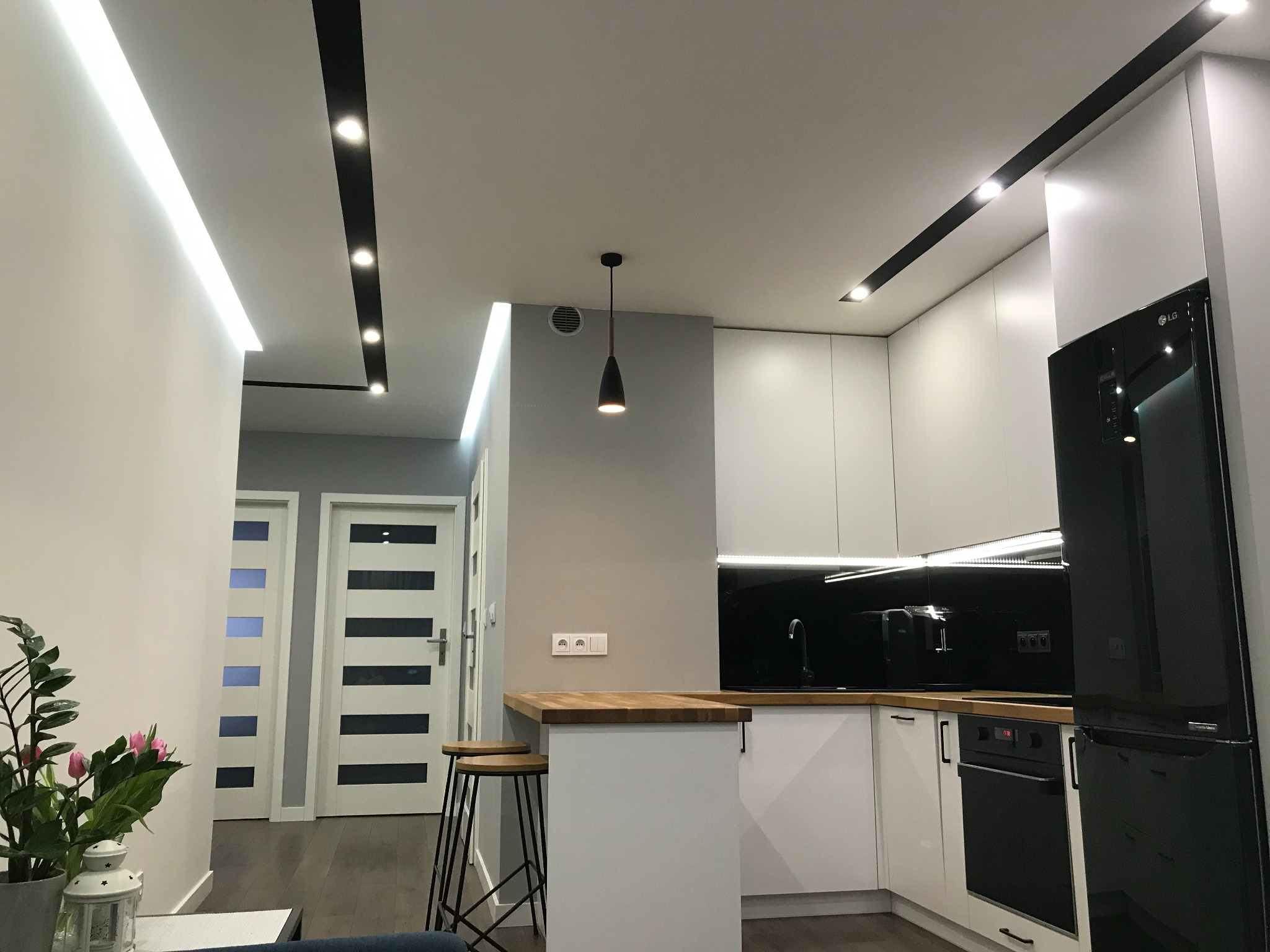 Nowoczesna kuchnia z białymi szafkami, czarnym panelem szklanym nad blatem, drewnianym blatem i czarną lodówką, oświetlona listwami LED i wiszącą lampą.