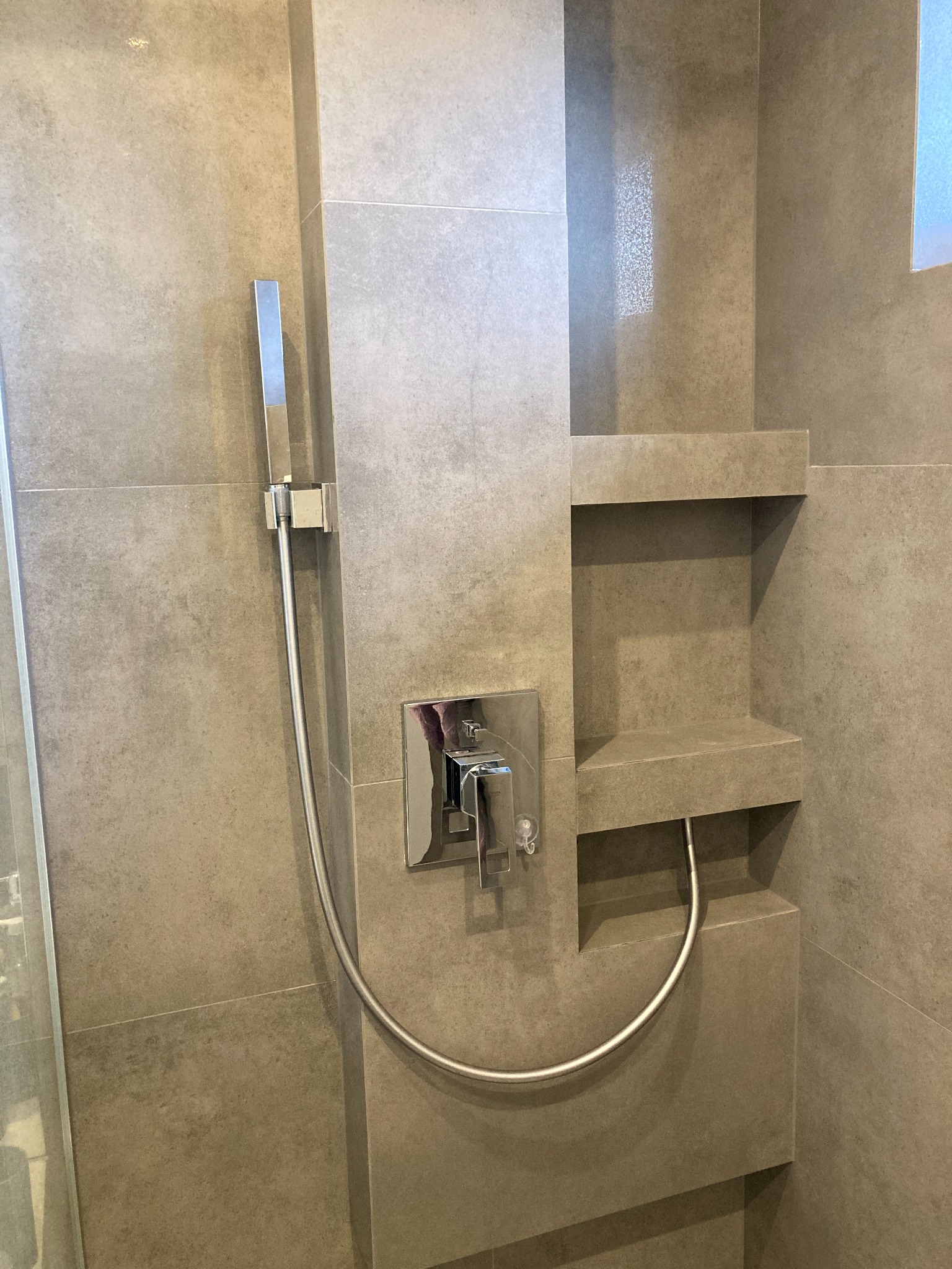 Nowoczesna kabina prysznicowa z szarymi, matowymi płytkami, wbudowanymi półkami i chromowaną armaturą, widoczna słuchawka prysznicowa i bateria.