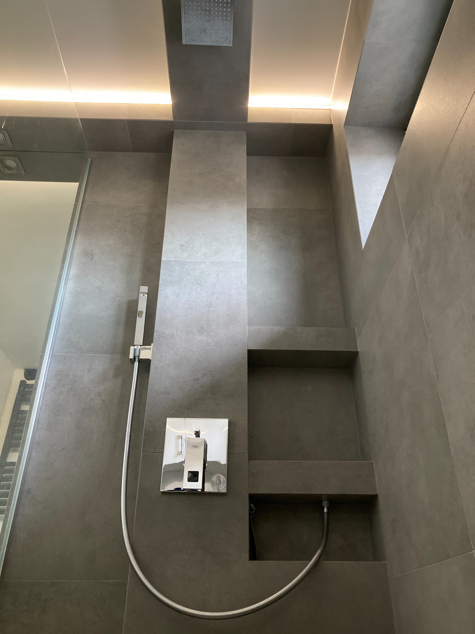 Nowoczesna kabina prysznicowa z szarymi płytkami, wbudowanymi półkami i oświetleniem LED, widoczna bateria Grohe.