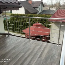 Błyszcząca, metalowa balustrada balkonowa w Skierniewicach, z widokiem na altanę i domy w tle.