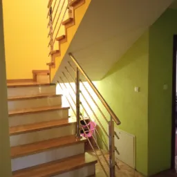 Schody z drewnianymi stopniami i metalową balustradą w Skierniewicach, oświetlone ciepłym światłem, na tle żółtej i zielonej ściany.