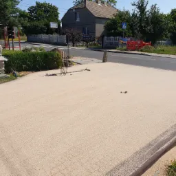 Przygotowany podkład piaskowy pod przyszłą nawierzchnię, widoczne narzędzia i materiały budowlane, w tle dom i ulica.