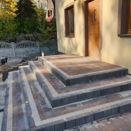 PRO BRUK Wojciech Zabiegała - Schody wejściowe z kostki brukowej w odcieniach szarości i brązu, prowadzące do drzwi domu. Widoczne detale ułożenia kostki i otoczenie budynku.