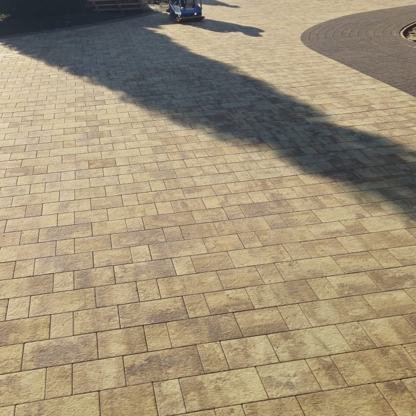 Beżowa kostka brukowa ułożona w regularny wzór, z widocznym cieniem drzewa i zagęszczarką w tle. Nowa nawierzchnia z kostki brukowej.