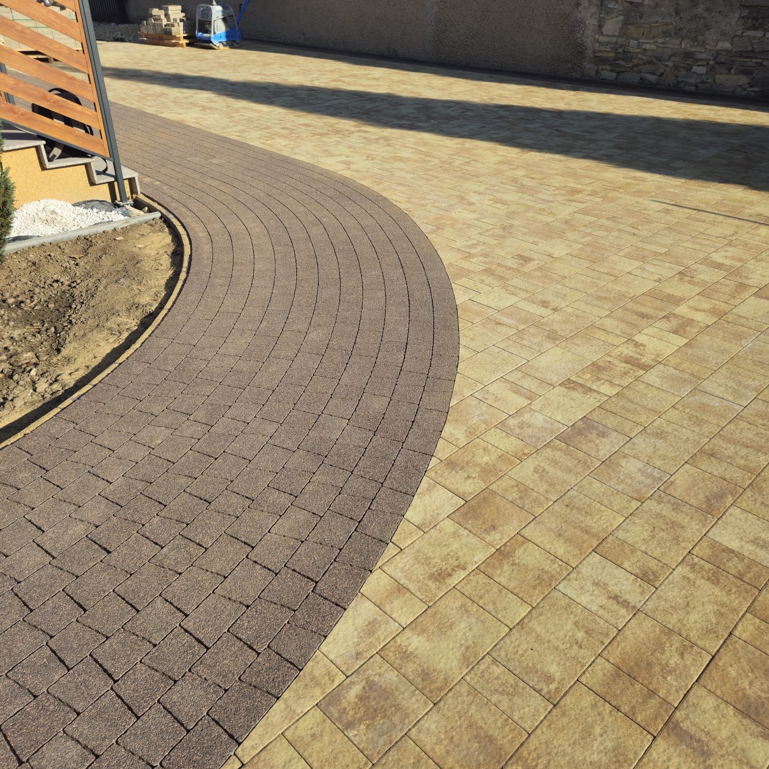 Ułożona kostka brukowa w dwóch odcieniach: brązowa w łuku i beżowa w prostokątnym wzorze. Widoczne schody z drewnianą barierką i paleta z kostką w tle.