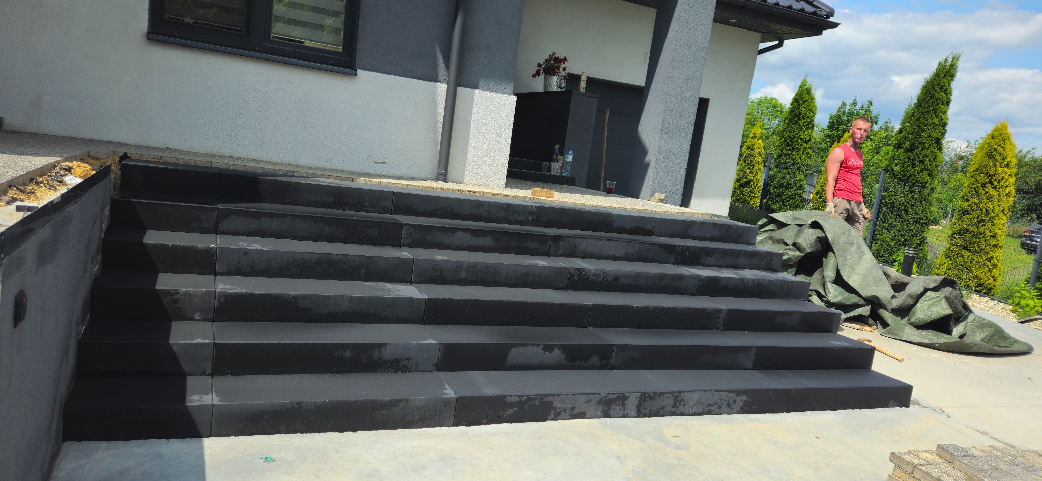 Nowoczesne, czarne schody wejściowe z kostki brukowej prowadzące do domu. Widoczny fragment elewacji i osoba w tle. Ujęcie z dołu, podkreślające solidność konstrukcji.