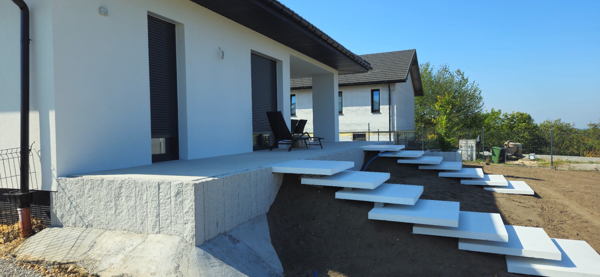 Nowoczesny taras z białymi, betonowymi stopniami prowadzącymi w dół zbocza. Widoczny fragment domu z szarymi roletami i kamiennym wykończeniem.