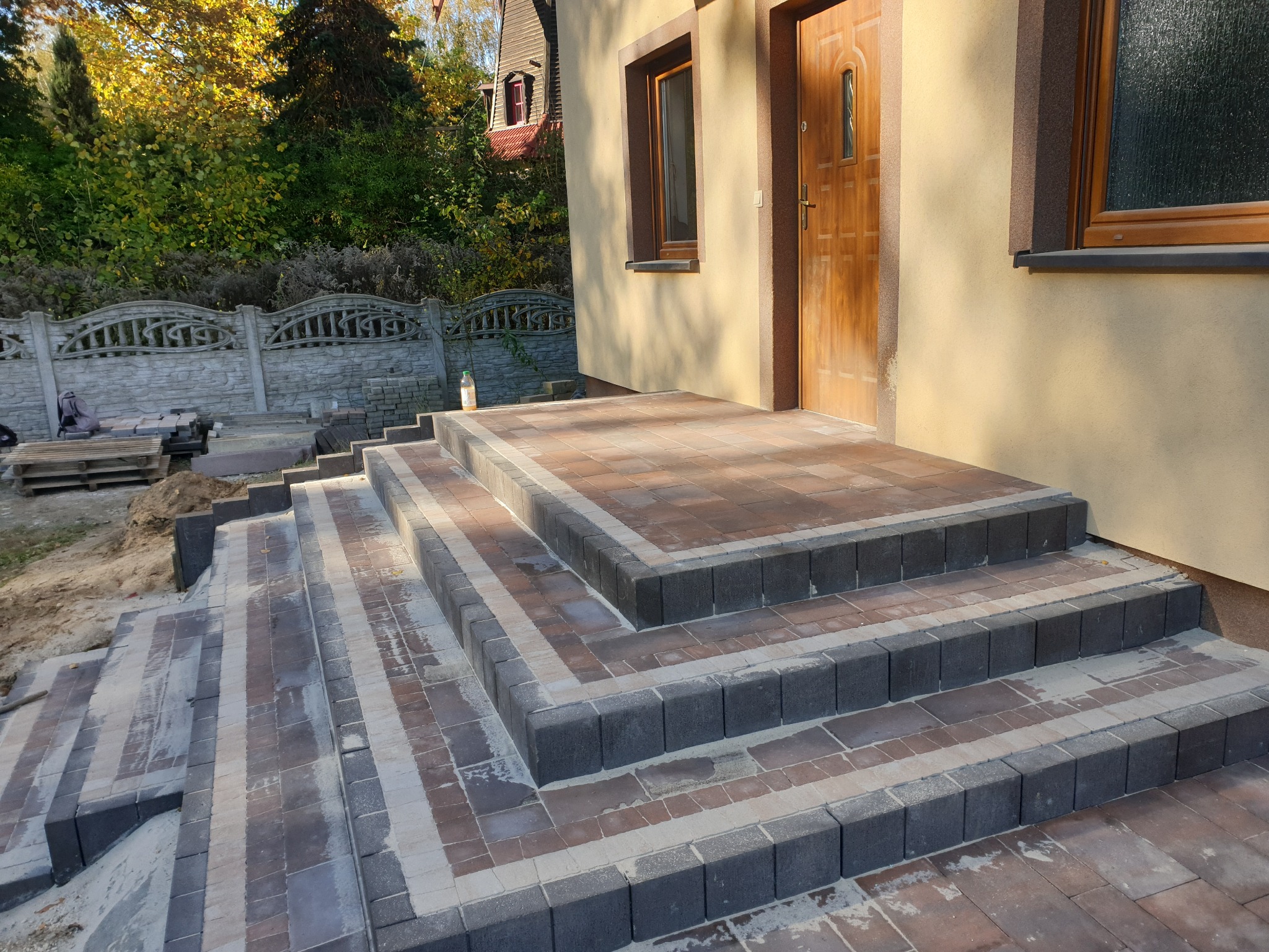 Schody wejściowe z kostki brukowej w odcieniach szarości i brązu, prowadzące do drzwi domu. Widoczne detale ułożenia kostki i otoczenie budynku.
