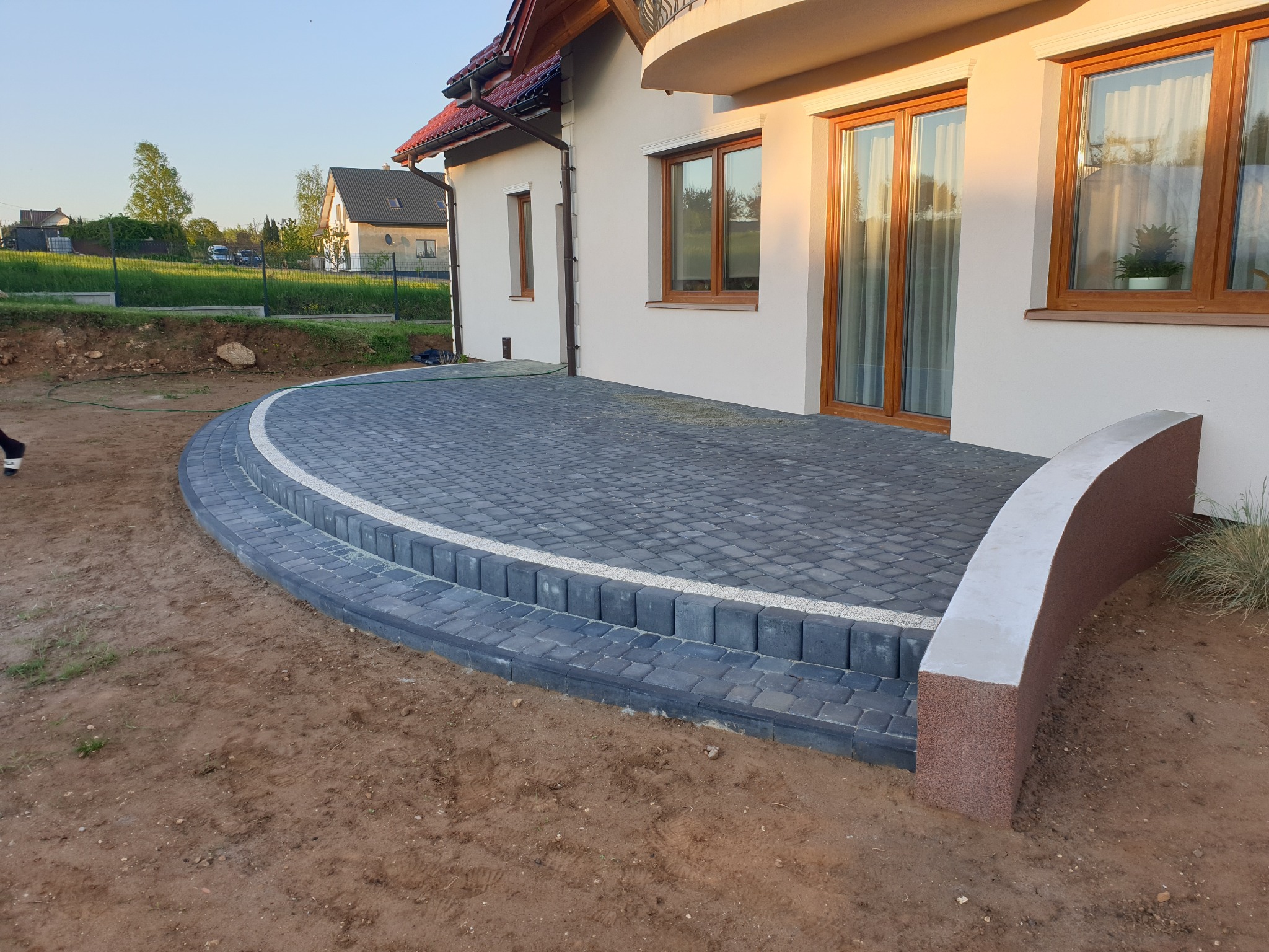 Elegancki taras z kostki brukowej w odcieniach szarości, z białym akcentem, przylegający do jasnego budynku z drewnianymi oknami. Widoczne detale wykończenia i otoczenie.