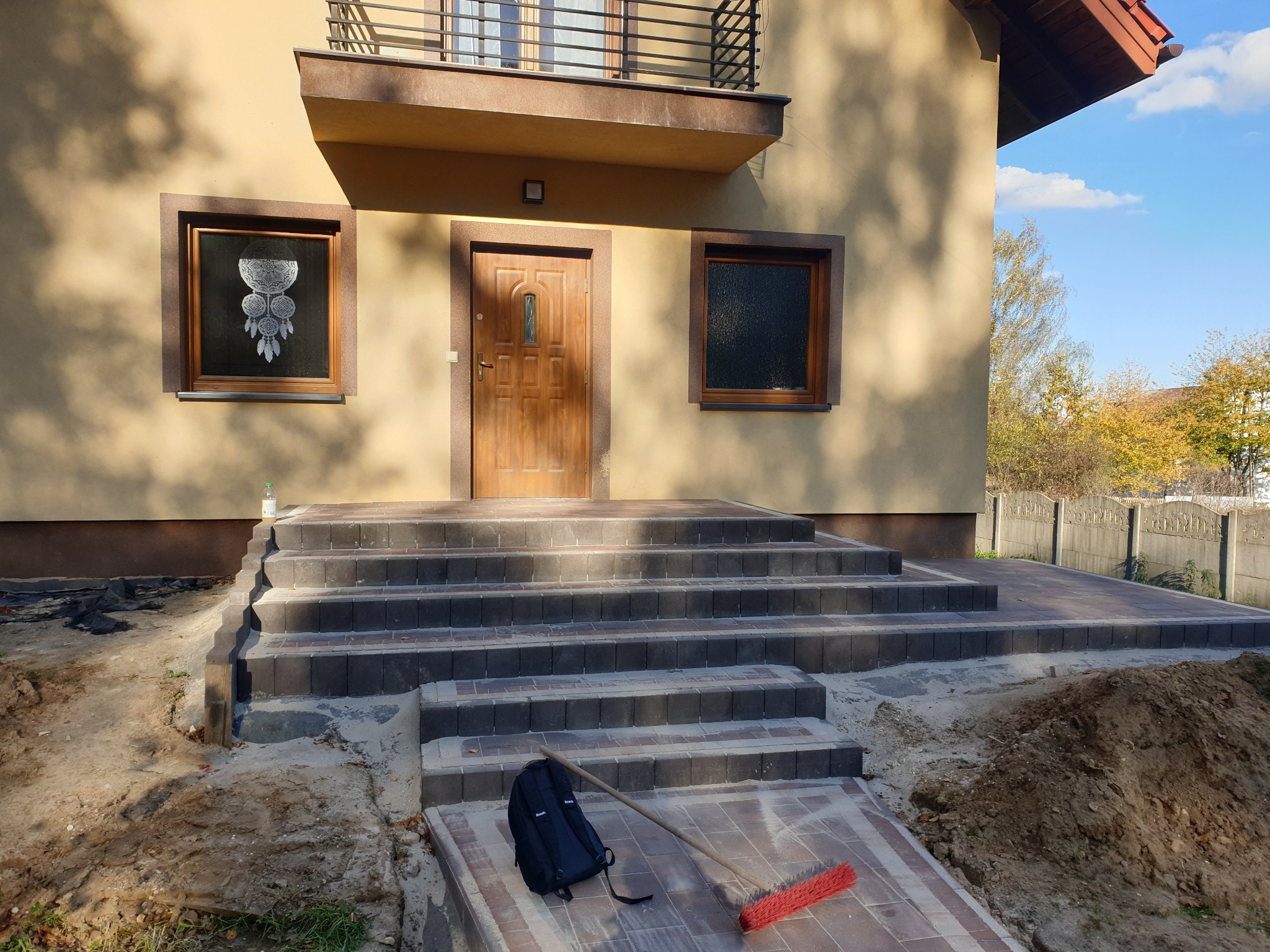 Schody wejściowe z kostki brukowej prowadzące do domu, z widocznym plecakiem i miotłą na pierwszym planie. Tło stanowi elewacja budynku w ciepłych barwach.