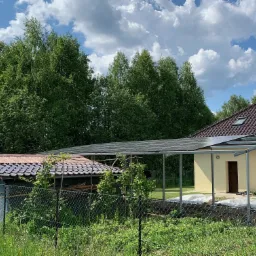 Panele słoneczne zamontowane na konstrukcji przy budynku jednorodzinnym z brązowym dachem, widok zza ogrodzenia z siatki, w tle zielone drzewa i błękitne niebo z chmurami.