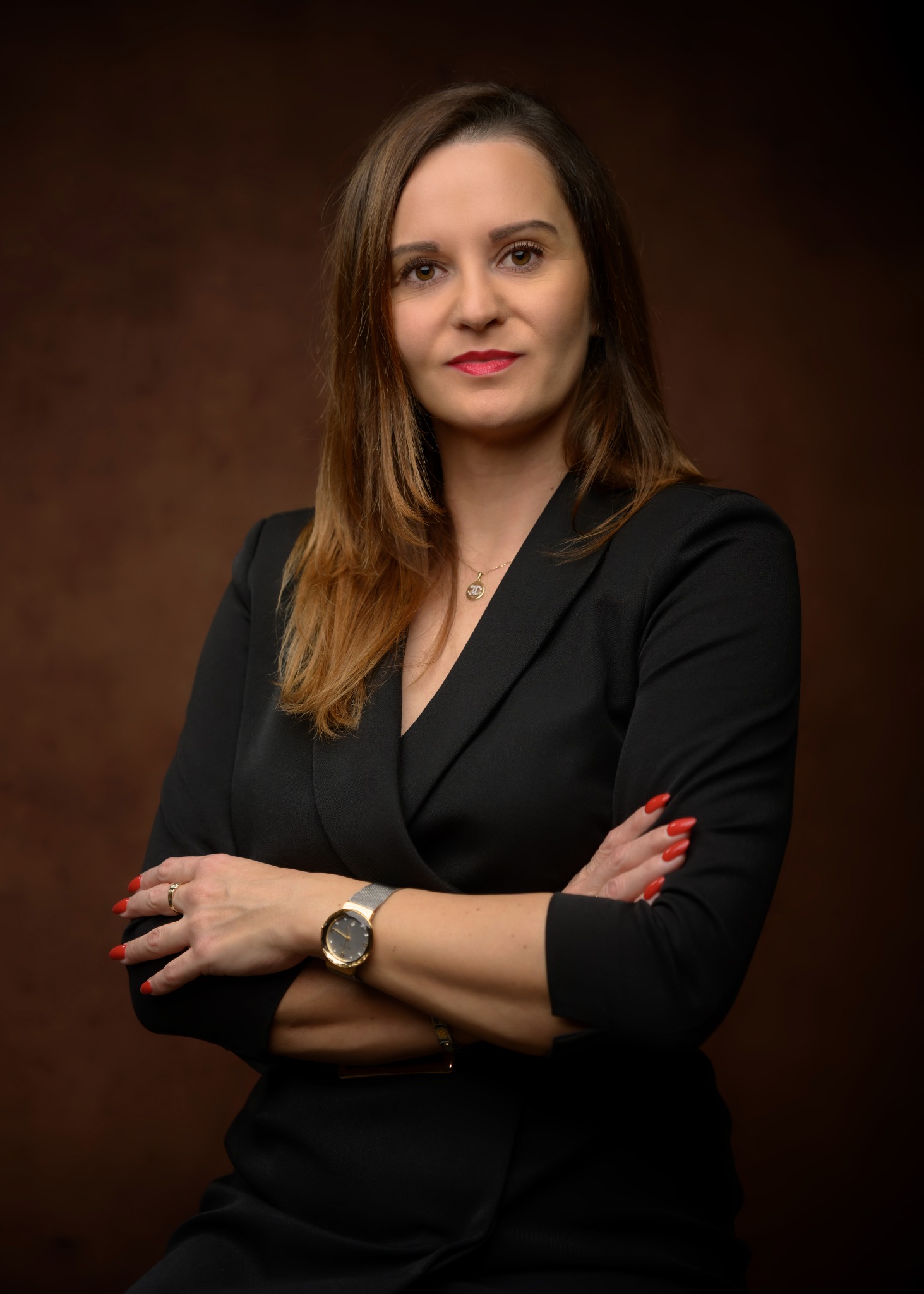 Portret kobiety w czarnym żakiecie na brązowym tle. Skrzyżowane ręce, czerwone paznokcie, złoty zegarek i delikatny naszyjnik. Profesjonalny wizerunek.
