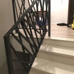 Czarna, geometryczna barierka schodowa z metalu, z ukośnymi prętami tworzącymi wzór krzyży, zamontowana przy białych schodach w jasnym wnętrzu.