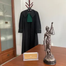 Wnętrze kancelarii: toga adwokacka na wieszaku, statuetka Temidy z wagą i mieczem, wizytówka na biurku z drewnianym blatem i przeszklona szafa na dokumenty w tle.