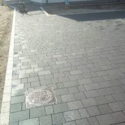 Ułożona szara kostka brukowa z widoczną studzienką kanalizacyjną, obrzeża wykończone jaśniejszą kostką. W tle biały budynek z oknem i ubijarka wibracyjna.