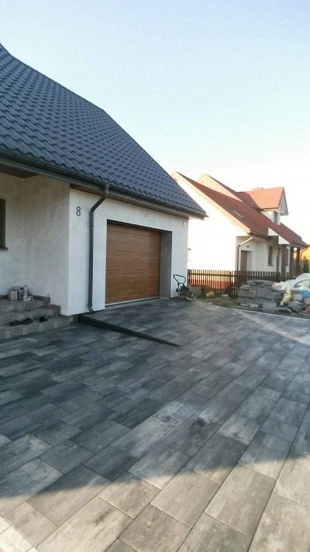 Ułożona szara kostka brukowa na podjeździe przed domem z brązową bramą garażową, w tle drugi dom z czerwonym dachem.