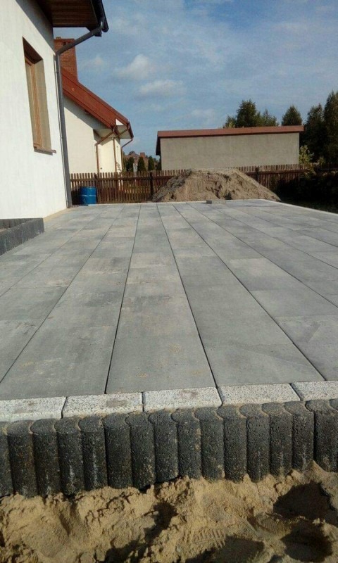 Szara, prostokątna kostka brukowa ułożona na posesji, obrzeża z ciemnych, okrągłych elementów, w tle dom jednorodzinny i sterta piasku.