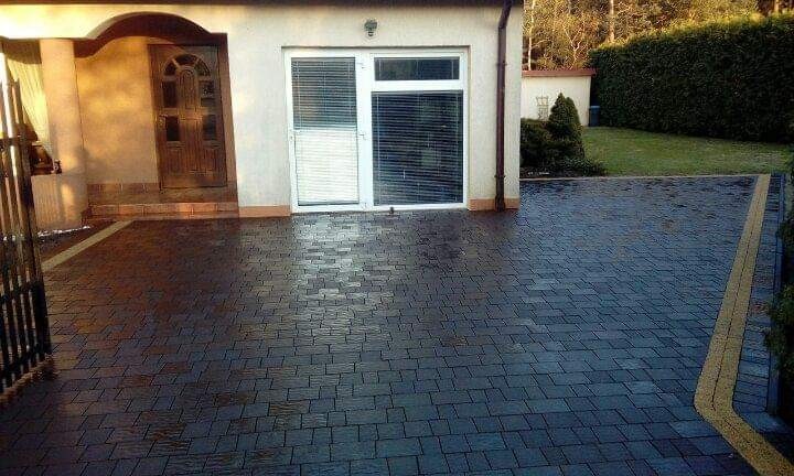Ułożona, ciemna kostka brukowa na podjeździe przed domem z bramą i drzwiami wejściowymi, widoczny żółty pasek oddzielający kostkę od trawnika.