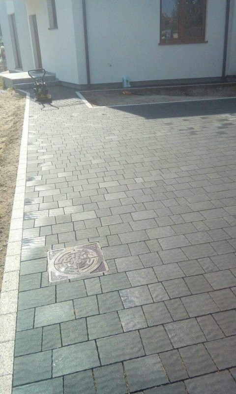 Ułożona szara kostka brukowa z widoczną studzienką kanalizacyjną, obrzeża wykończone jaśniejszą kostką. W tle biały budynek z oknem i ubijarka wibracyjna.