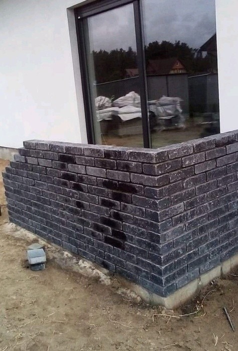 Fragment elewacji budynku w trakcie wykańczania kostką brukową w kolorze grafitowym, widoczne niedokończone rzędy i zapas kostki obok, okno z odbiciem otoczenia w tle.