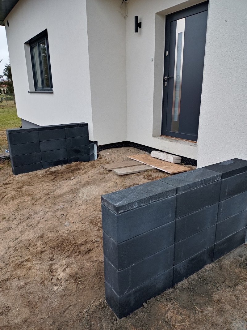 Częściowo ułożony murek z ciemnej kostki brukowej przed wejściem do nowoczesnego domu z szarą stolarką okienną i drzwiową, widoczne deski i piasek.