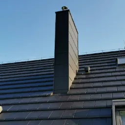 Nowoczesny, ciemny dach z ceramicznej dachówki karpiówki, z czarnym kominem obłożonym blachą na tle błękitnego nieba, zabezpieczenia przeciwśniegowe wzdłuż kalenicy.