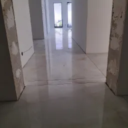 Korytarz w trakcie remontu, widoczne świeżo położone, błyszczące, beżowe płytki podłogowe imitujące marmur oraz surowe, nieotynkowane futryny drzwiowe.