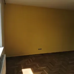 Pomalowany na żółto pokój z parkietem w jodełkę; widoczne gniazdka elektryczne w ścianie i grzejnik pod oknem.