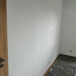 Świeżo pomalowane na biało wnętrze pokoju z drewnianą listwą przypodłogową i zabezpieczoną folią podłogą, widoczne otwory po gniazdkach.