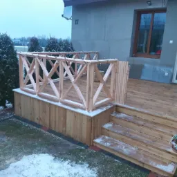 Drewniany taras z balustradą, schodami i oświetleniem zewnętrznym przy domu w zimowej scenerii, częściowo pokryty śniegiem.