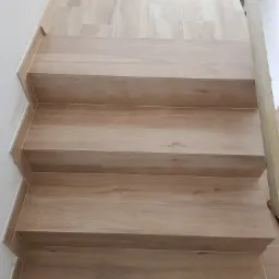 Schody wyłożone płytkami imitującymi drewno, z widocznymi fugami i listwami wykończeniowymi, częściowo zakryte przez drewniany element konstrukcyjny i różową szmatkę.