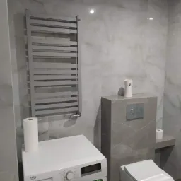 Wykończona łazienka z szarymi marmurkowymi płytkami, grzejnikiem drabinkowym, toaletą podwieszaną, białą pralką Beko i rolką papieru toaletowego na pralce.