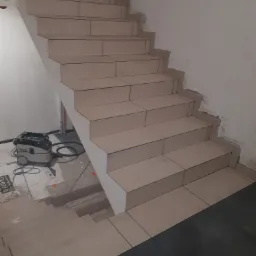 Wyłożone płytkami schody wewnętrzne w trakcie prac wykończeniowych, widoczny odkurzacz przemysłowy i częściowo wykończona podłoga.