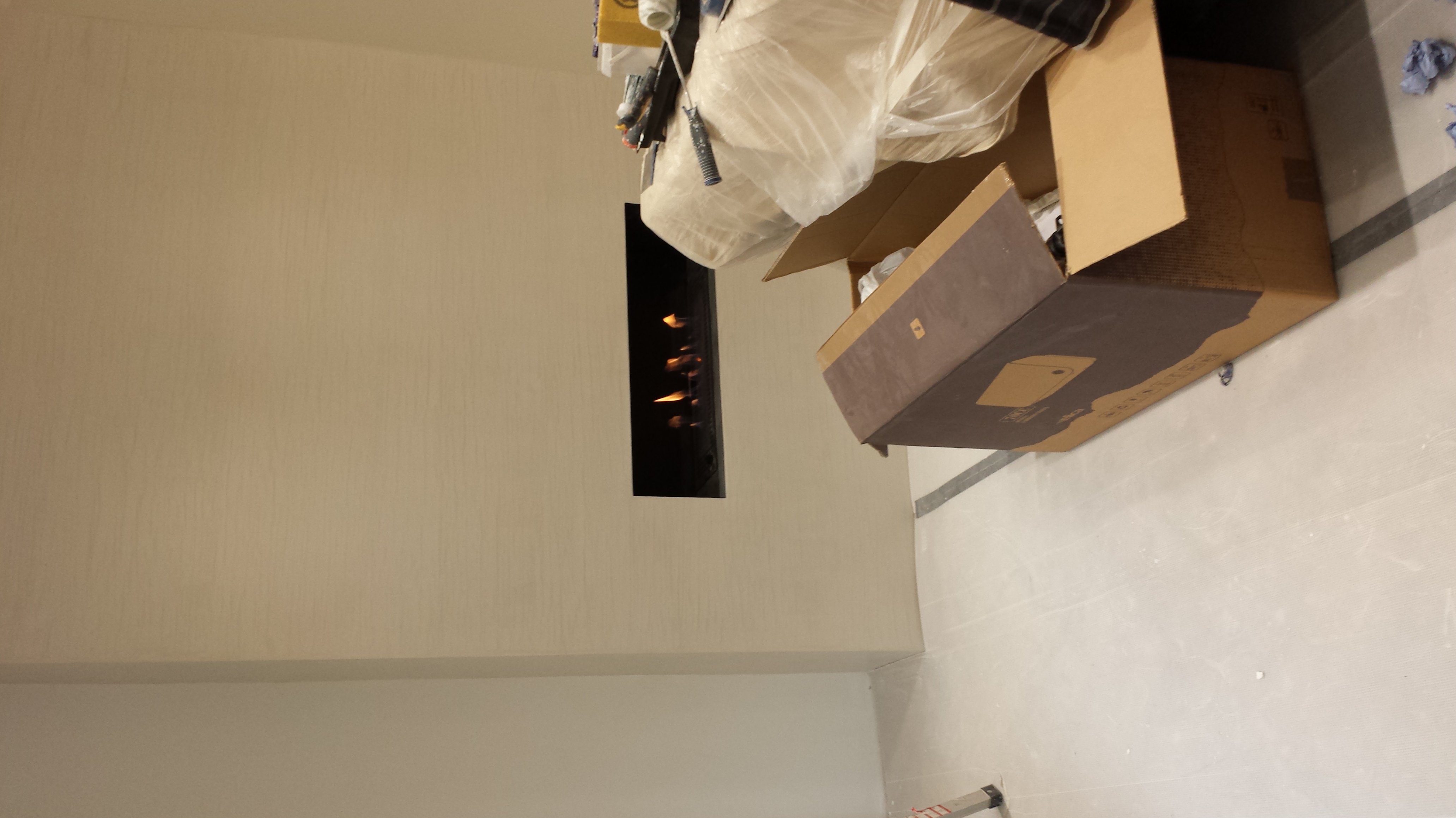 Ściana po nałożeniu gładzi, z wbudowanym kominkiem elektrycznym, narzędzia malarskie pod folią ochronną i rozpakowane opakowanie produktu na podłodze.