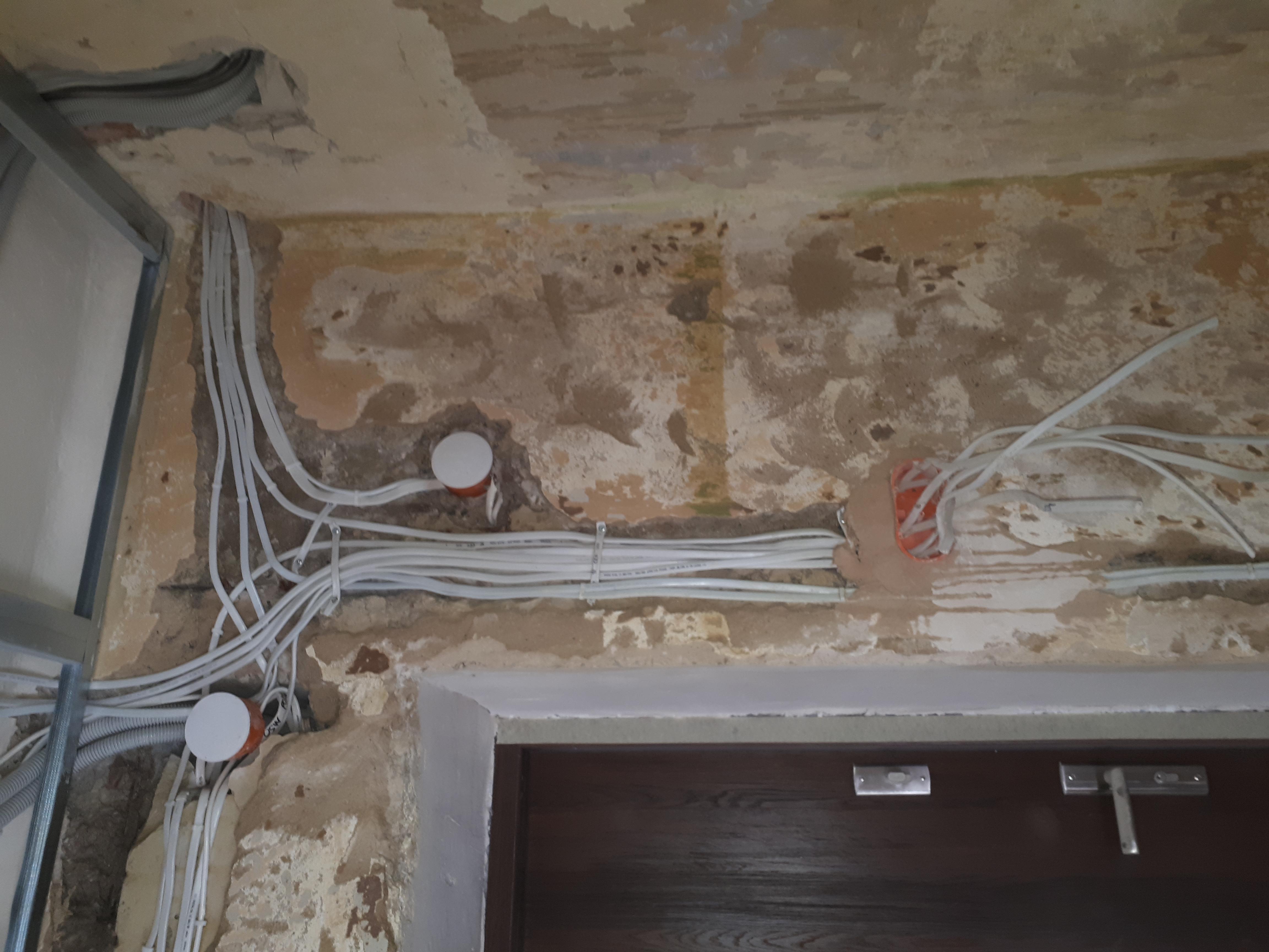 Instalacja elektryczna w trakcie remontu, widoczne kable w peszlach i puszki elektryczne w ścianie z odsłoniętym murem, fragment metalowej konstrukcji sufitu podwieszanego.