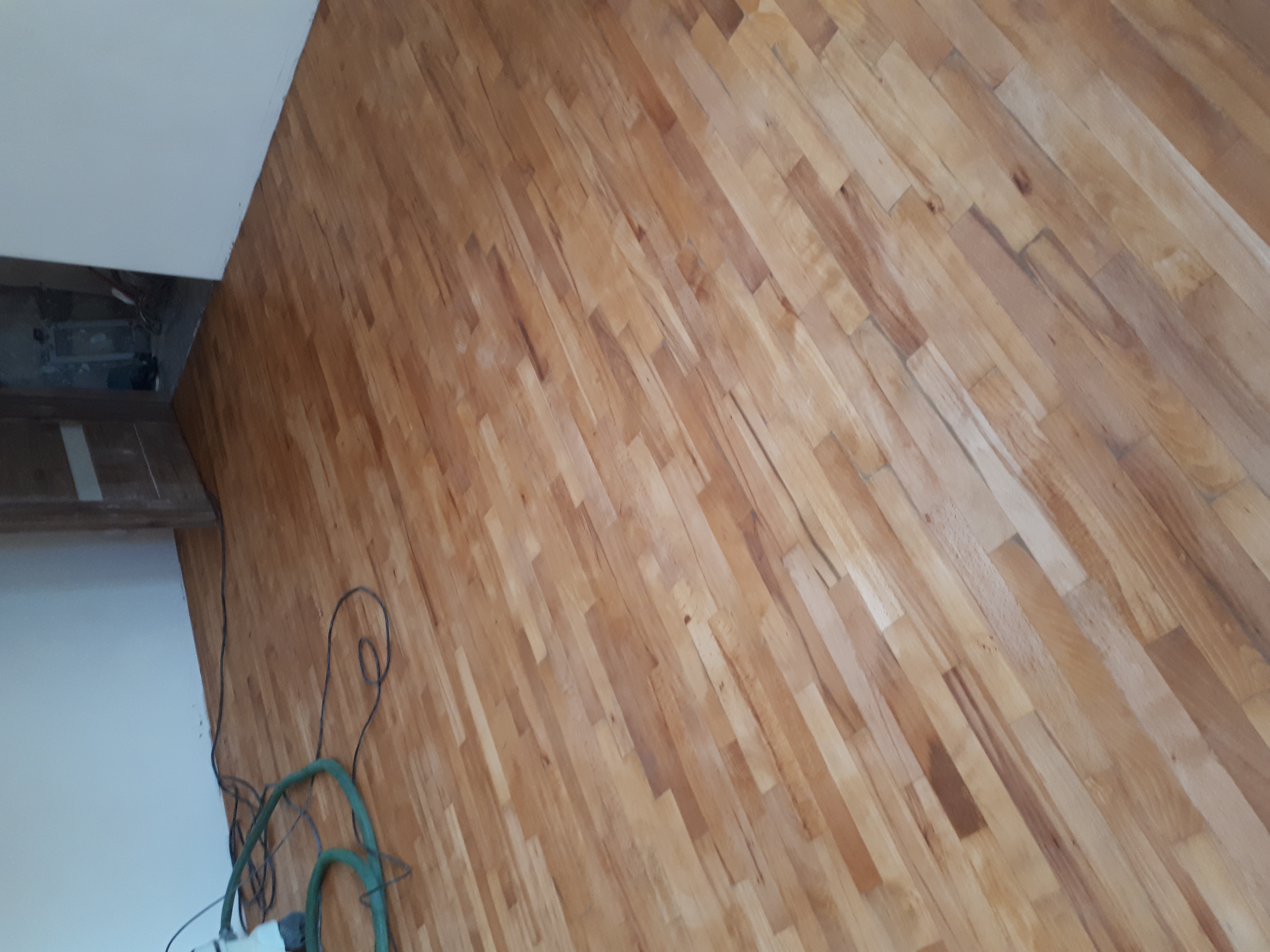 Ułożona, lakierowana podłoga z parkietu drewnianego w odcieniach jasnego brązu, widoczne przewody i fragment odkurzacza przy ścianie.