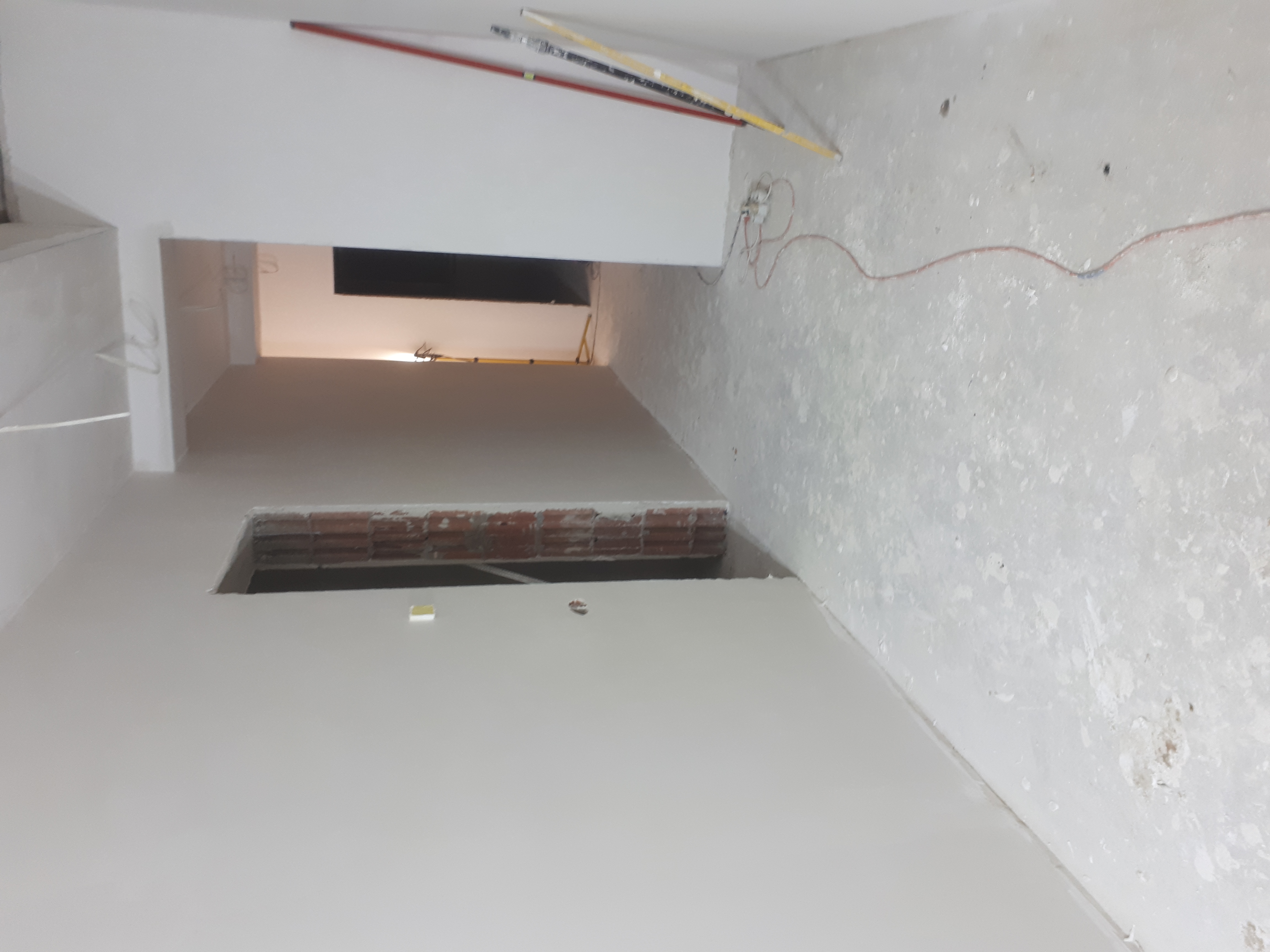 Wnętrze w trakcie remontu z surowymi ścianami i podłogą, widoczne przewody elektryczne i fragment ściany z cegieł, przygotowane do dalszych prac wykończeniowych.