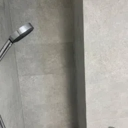 Szczegółowe ujęcie kabiny prysznicowej z szarymi płytkami imitującymi kamień, widoczna słuchawka prysznicowa i szklana półka z czerwonym pojemnikiem.