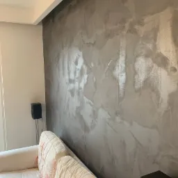 Ściana w salonie pokryta dekoracyjną, szarą farbą imitującą marmur, z widoczną fakturą i refleksami światła, obok stoi sofa w kwiaty i czarny głośnik.