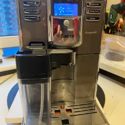 Automatyczny ekspres do kawy Saeco Incanto z wyświetlaczem wskazującym tryb czyszczenia, widoczny pojemnik na mleko, w tle okno i pojemnik z ziarnami kawy.