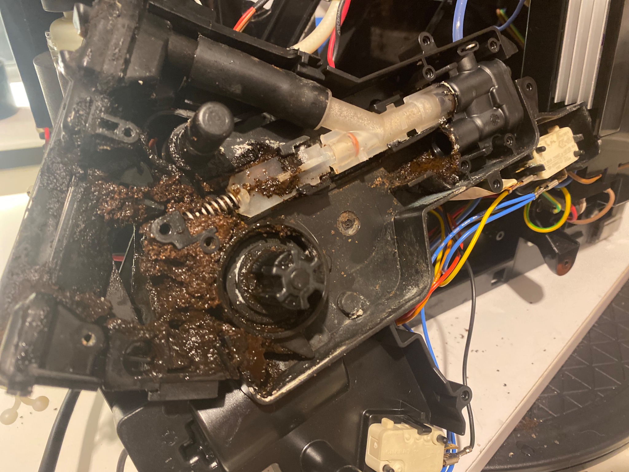 Wnętrze zepsutego ekspresu do kawy, widoczne elementy mechaniczne pokryte resztkami kawy i osadami, przewody elektryczne w tle.