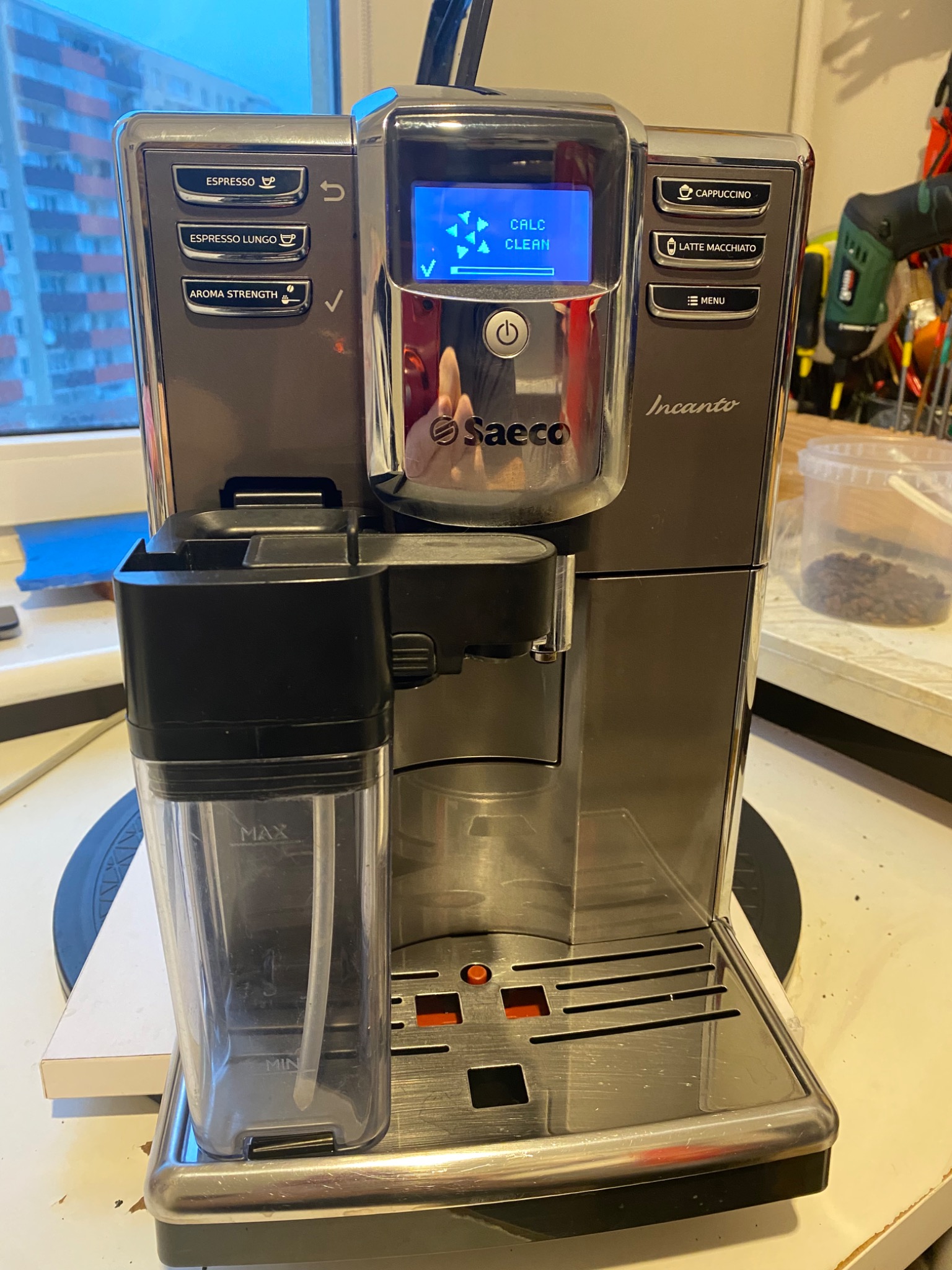 Automatyczny ekspres do kawy Saeco Incanto z wyświetlaczem wskazującym tryb czyszczenia, widoczny pojemnik na mleko, w tle okno i pojemnik z ziarnami kawy.