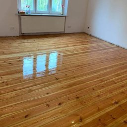 DFParkiet - Błyszcząca, świeżo polakierowana podłoga z desek w jasnym odcieniu drewna, z widocznym odbiciem okna, w pustym pokoju z białymi ścianami i grzejnikiem pod oknem.