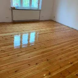 Błyszcząca, świeżo polakierowana podłoga z desek w jasnym odcieniu drewna, z widocznym odbiciem okna, w pustym pokoju z białymi ścianami i grzejnikiem pod oknem.