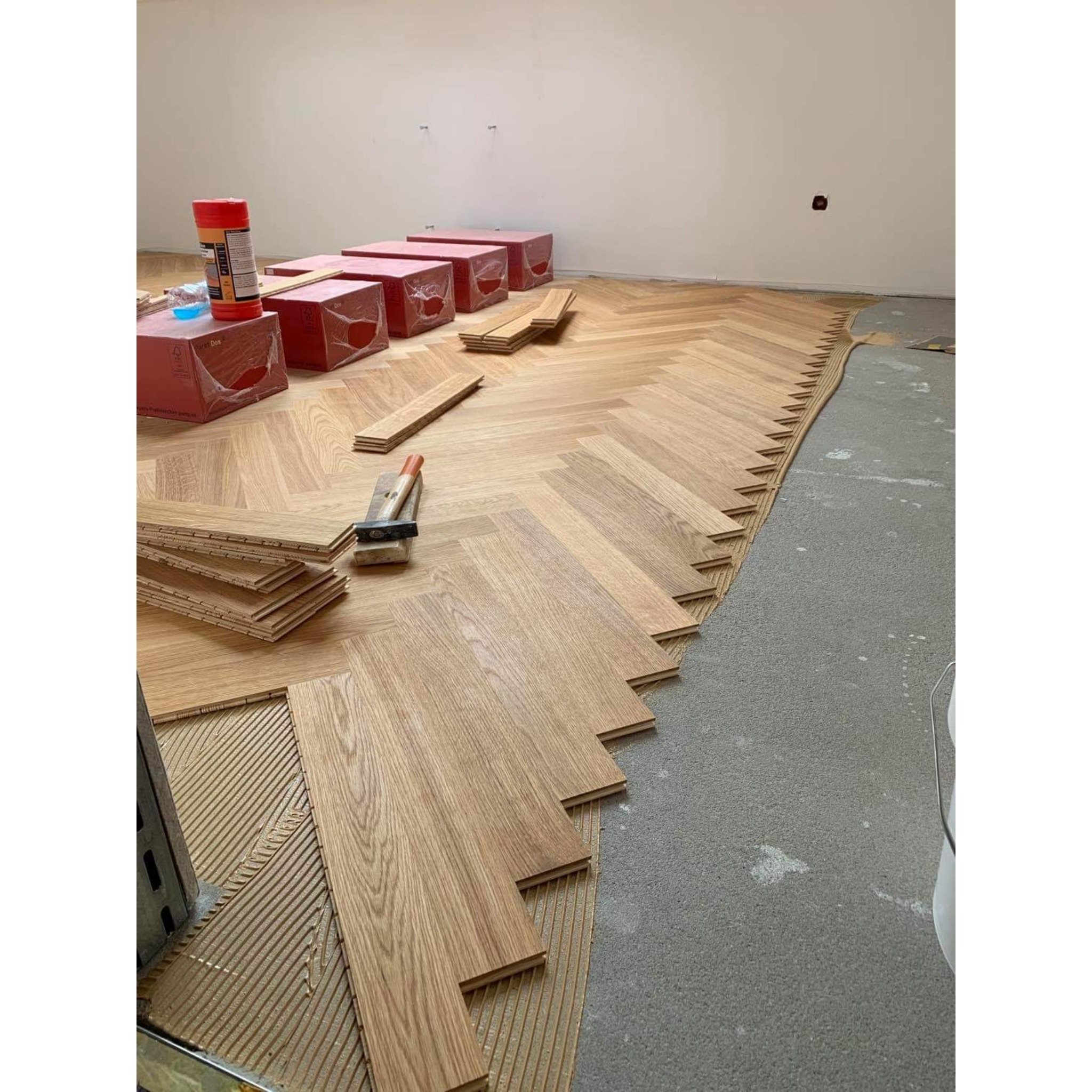 Układanie parkietu w jodełkę francuską, widoczne paczki z panelami, klej, narzędzia i fragment surowego betonu