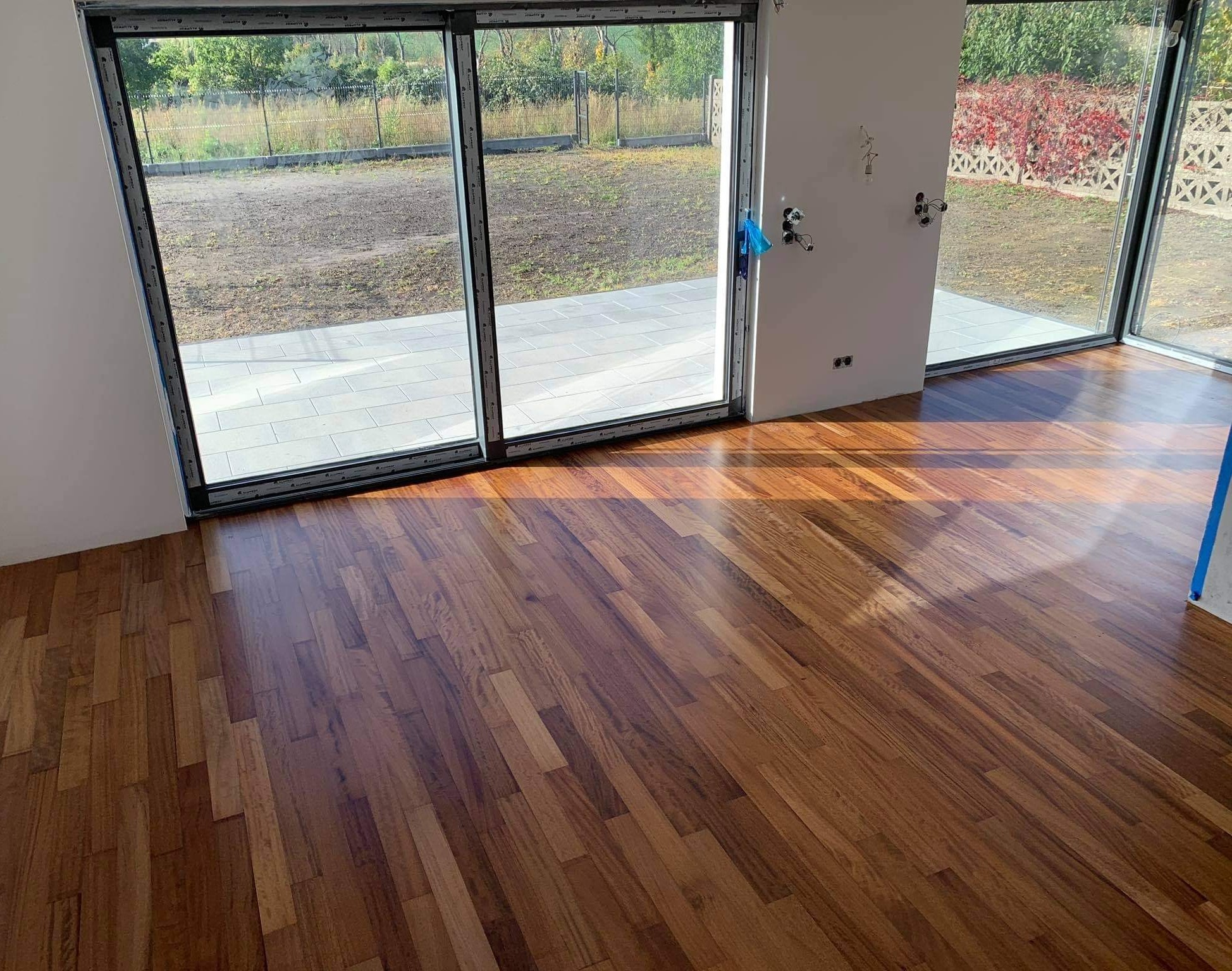 Świeżo położony, błyszczący parkiet z egzotycznego drewna w jasnym pomieszczeniu z dużymi przesuwnymi oknami tarasowymi i przygotowaną instalacją wodną na ścianie.