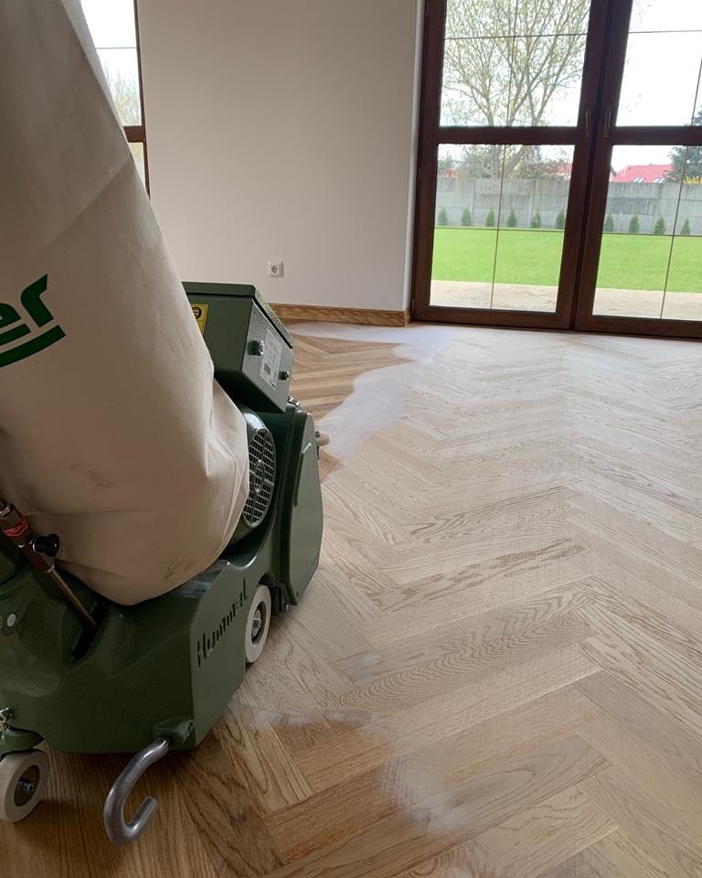 Parkiet w jodełkę poddawany obróbce cykliniarką, widoczny efekt rozjaśnienia drewna, duże okno z widokiem na trawnik w tle.