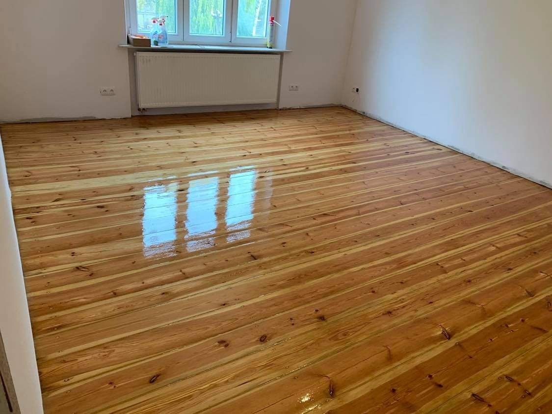 Błyszcząca, świeżo polakierowana podłoga z desek w jasnym odcieniu drewna, z widocznym odbiciem okna, w pustym pokoju z białymi ścianami i grzejnikiem pod oknem.