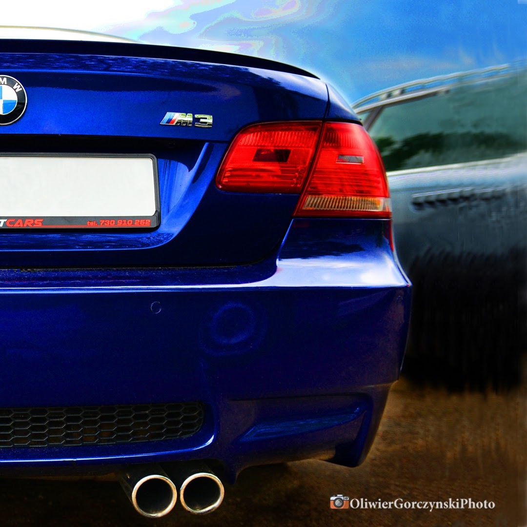 Tył sportowego, granatowego samochodu BMW M3 z widocznymi podwójnymi rurami wydechowymi i tablicą rejestracyjną z ramką reklamową